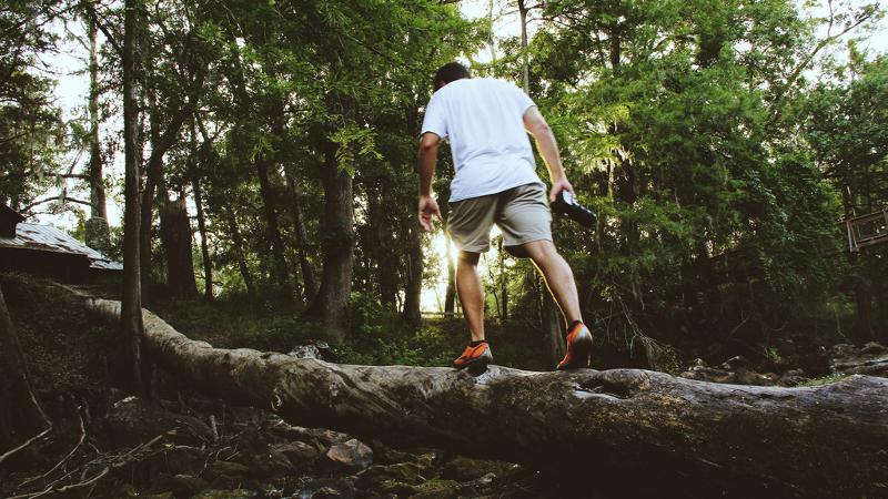 a man in a white shirt and shorts climbs on downed a tree branch carrying a water bottle